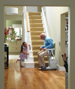 Person sitting on a stair lift at base of stairs speaking with a young child.