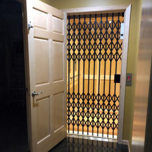 White door opening into a traditional home elevator with scissor gate and wooden shaker panels.