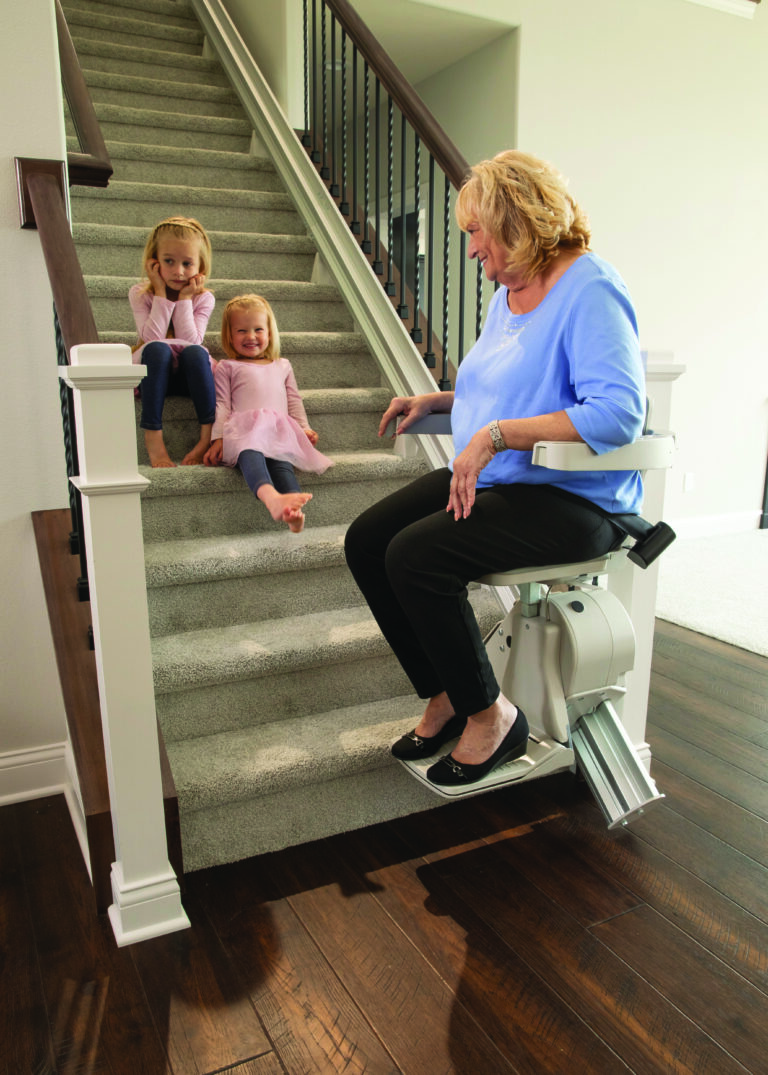 Woman sitting in stair lift chair with two children sitting on the stairs