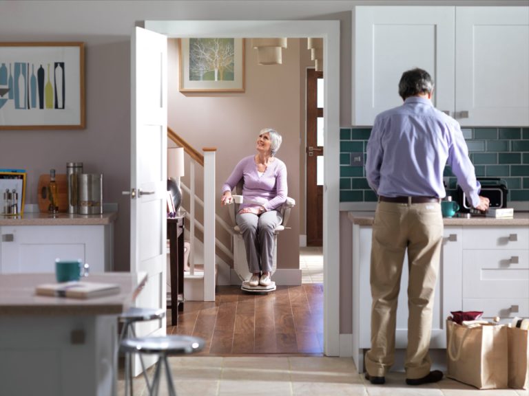 Woman sitting in stair lift chair through a doorway from a kitchen with man facing away in foreground