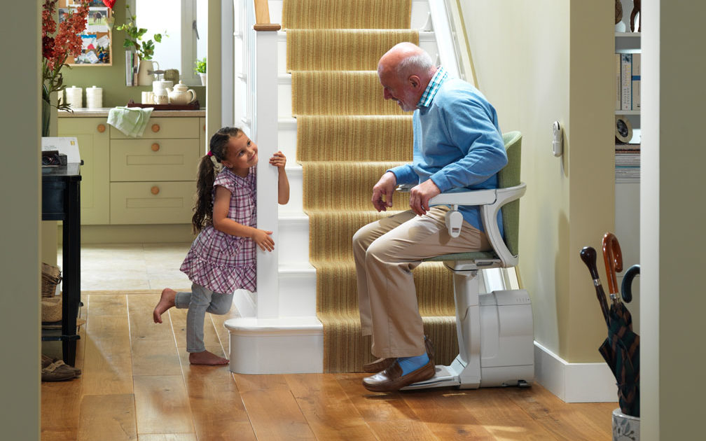 Man sitting in stair lift at base of stairs talking to young child. 