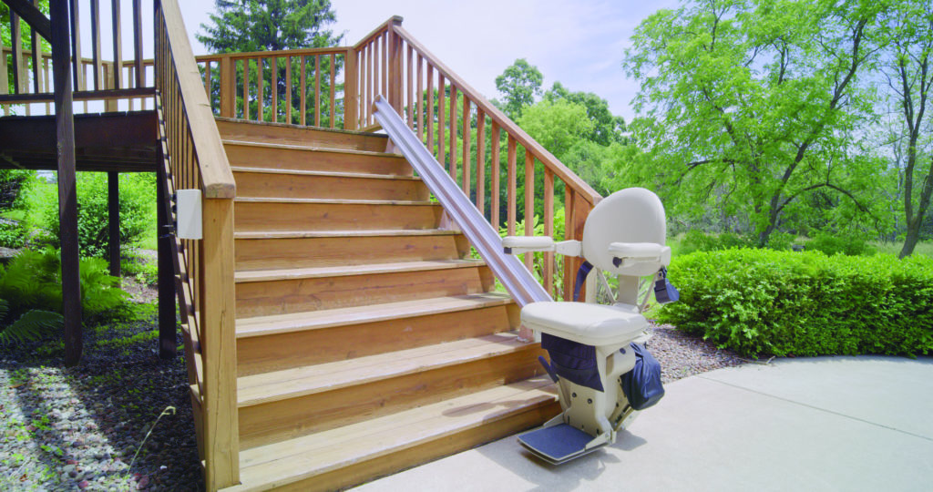 Outdoor stairglide with chair unfolded at base of a deck staircase on a sunny day.