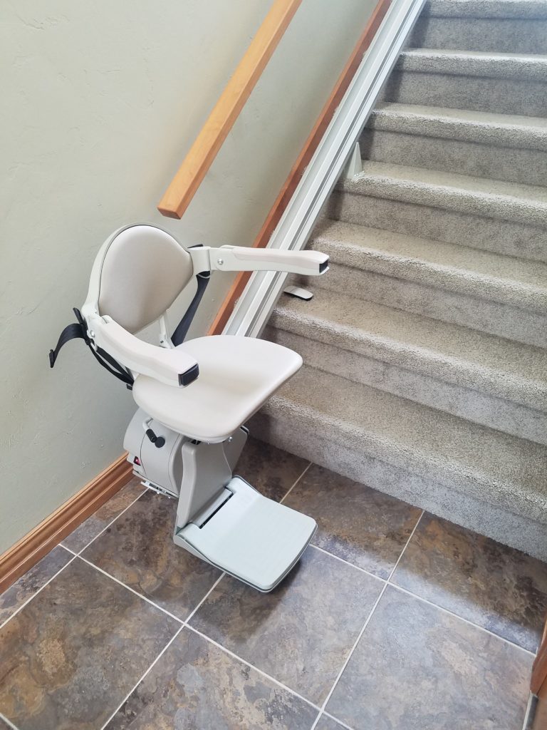 Used stairglide image installed in a residence. Stairglide is at base of stairs with chair, footrest and arms folded down ready for use.