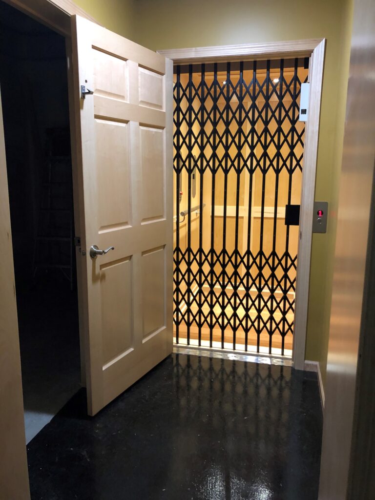 Traditional Home Elevator with raised wooden panels and a scissor gate door. The exterior door is open showing the scissor gate and paneling.