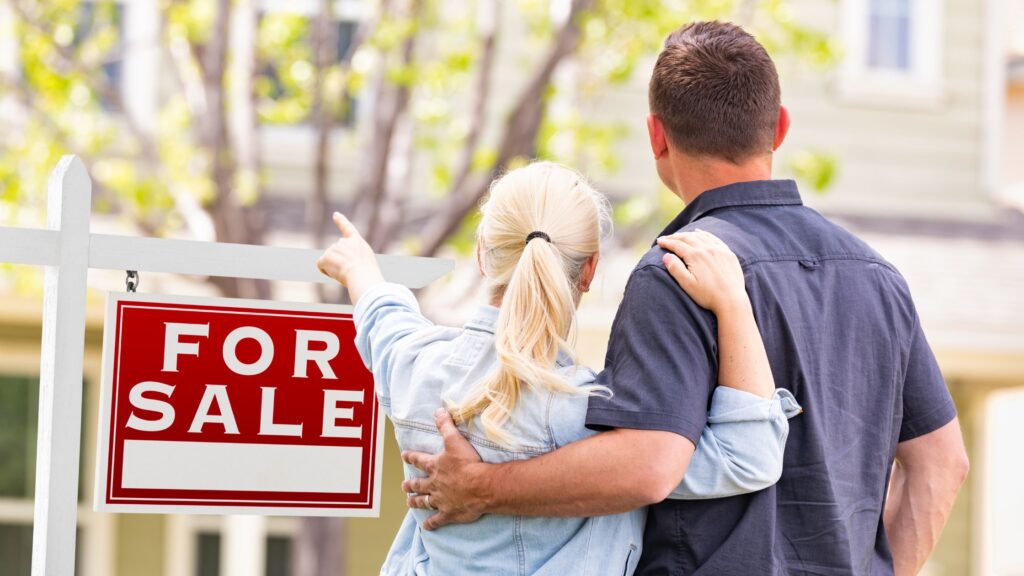 A man and woman embrace as they look at a home in the distance hidden behind a red for sale sign and a tree with fresh green buds.