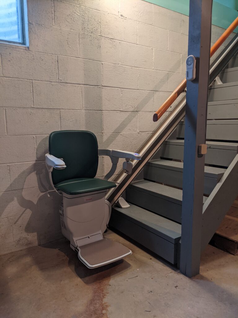 Green stair lift at the base of basement stairs with chair, arms and footrest in the down position.