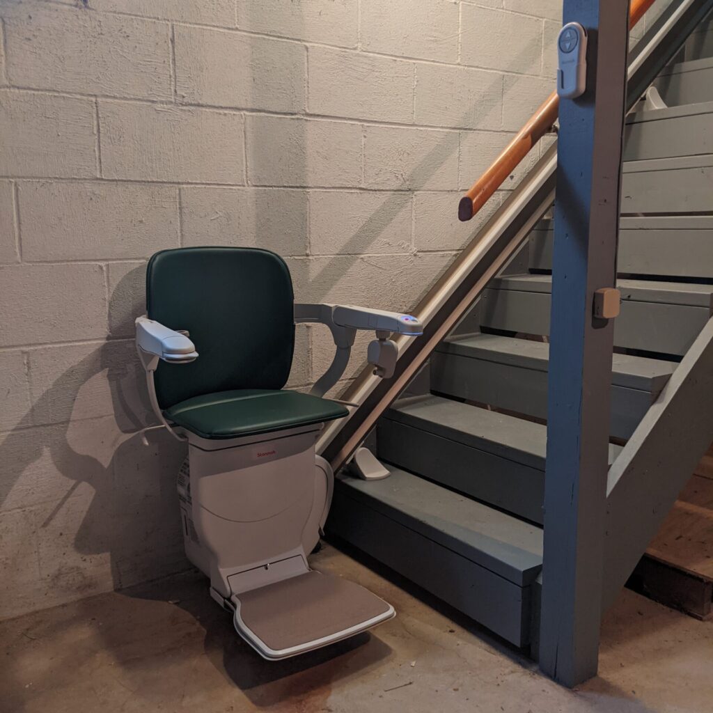 Green stair lift at the base of basement stairs with chair, arms and footrest in the down position.