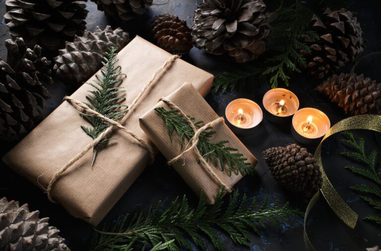 boxes wrapped in brown paper tied with twine and decorated with evergreen leaves next to lighted votive candles and encircled by pine cones