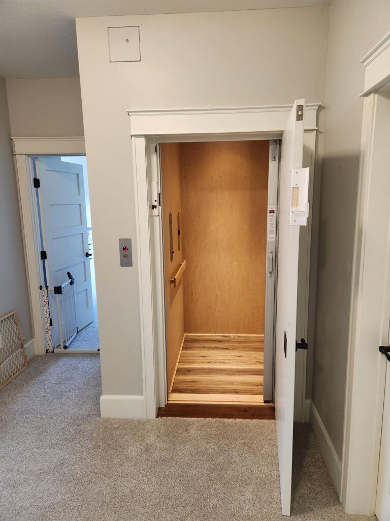 A wood-paneled home elevator with a swing-style landing door opens into a carpeted home interior.