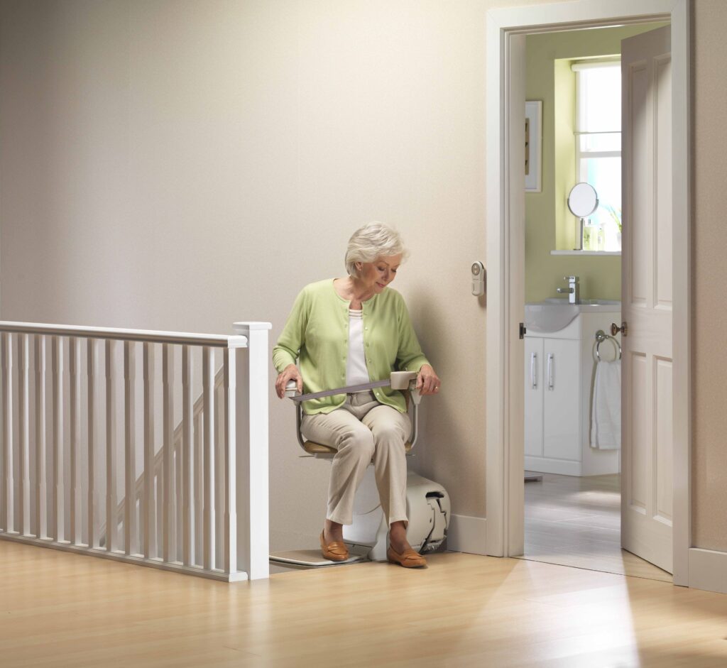 A woman sitting on a stair lift at the top of a staircase, safely buckled into the chair, swiveled toward the landing for a secure exit.