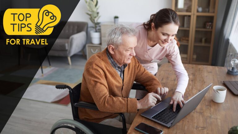 An older man in a wheelchair sits at a table with a laptop, while a smiling caregiver leans over to assist him.