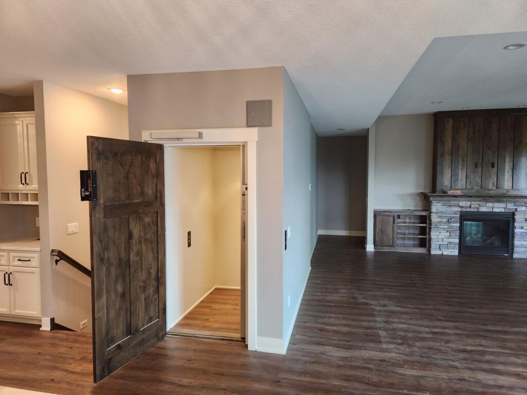 Residential home elevator with a custom wood door, installed in a modern living space with hardwood floors and stone fireplace.