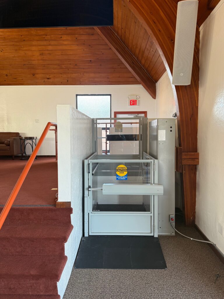 Vertical platform lift installed in a church, providing accessible entry to a raised stage with wood beam ceiling and carpeted flooring.