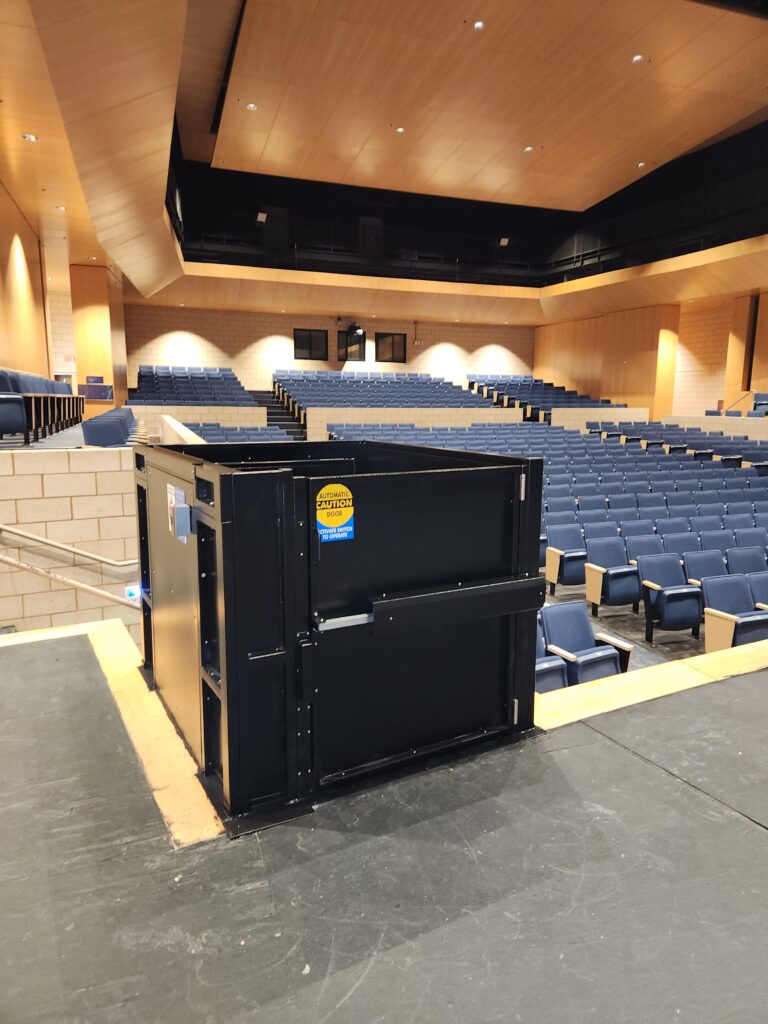 Black vertical platform lift installed on auditorium stage to provide wheelchair accessibility between stage and seating area in a modern high school theater.