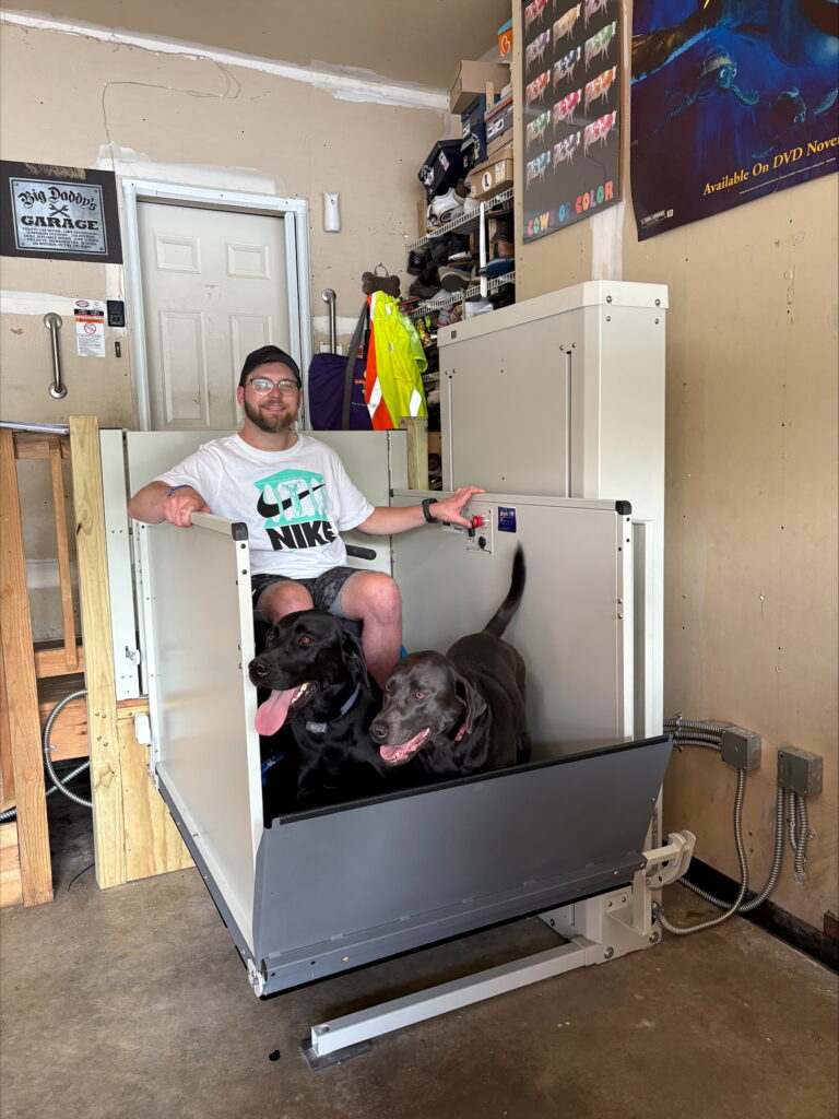 A man seated in a wheelchair smiles at the camera while riding his Vertical Platform Lift with his two dogs